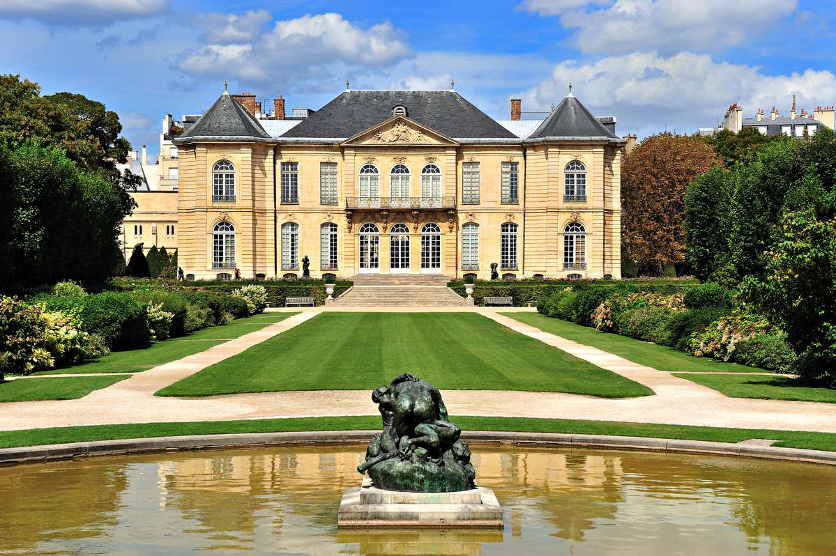 Musée Rodin vu de face avec un jardin et des allées, fontaine avec une statue au milieu au premier plan