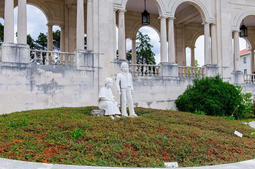 Duas estátuas de crianças em um jardim. A menina, sentada, segura um terço