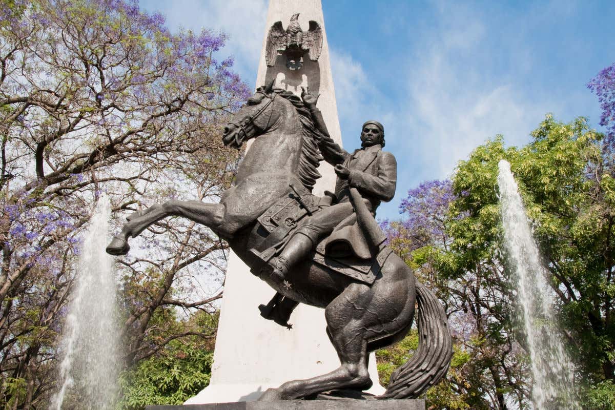 Escultura ecuestre realizada en bronce de José María Morelos frente a un obelisco de piedra blanca, dos chorros de agua procedentes de una fuente y varios árboles.