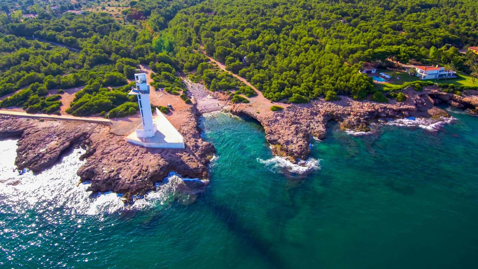 Aerial shot over a rocky coast with a small white lighthouse surrounded by many green trees