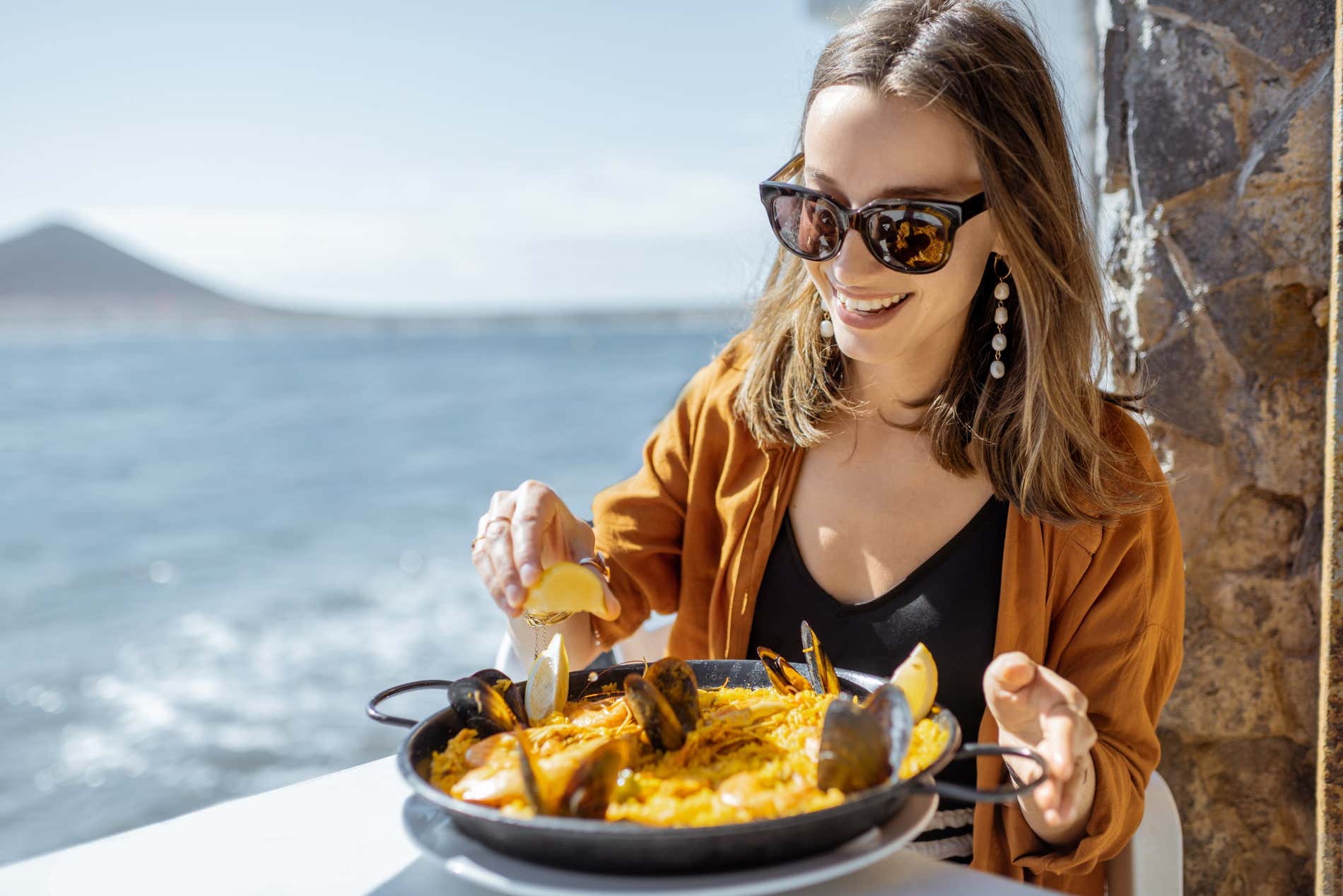 A woman squeezes a slice of lemon over an individual sea food paella at a table next to the sea in the Region of Valencia 