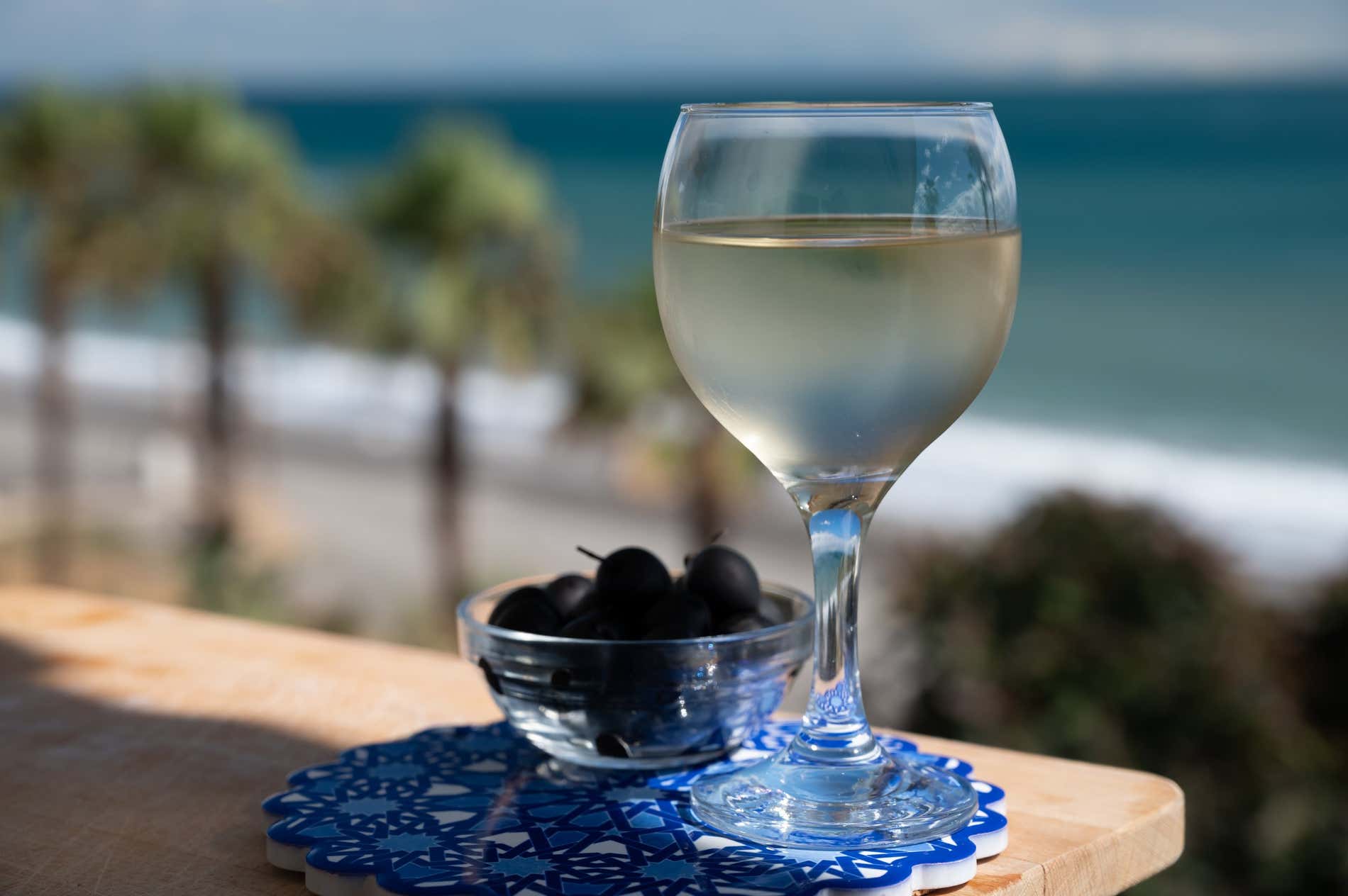A glass of pale wine next to a glass dish of black olives with a blurred view of palm trees and the sea behind