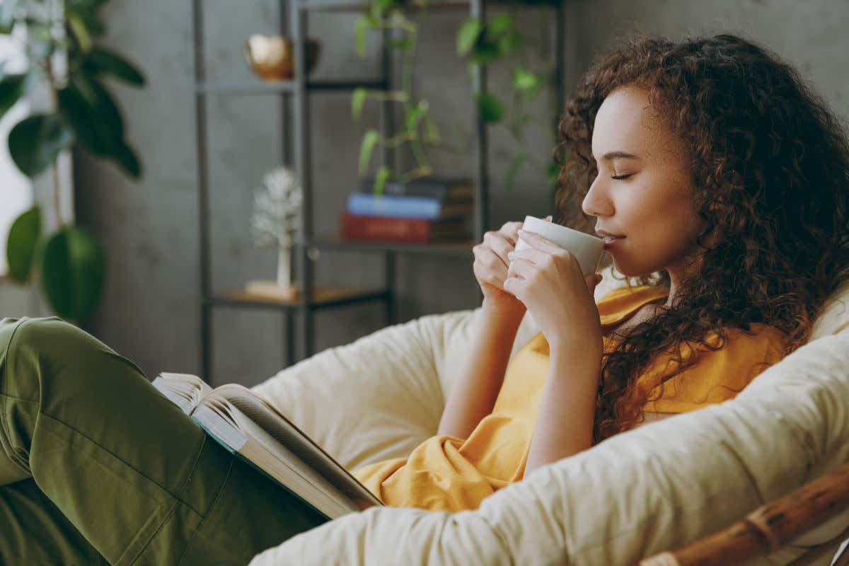 Una mujer de pelo rizado y piel oscura saboreando una taza de café en el sofá de su casa mientras lee un libro