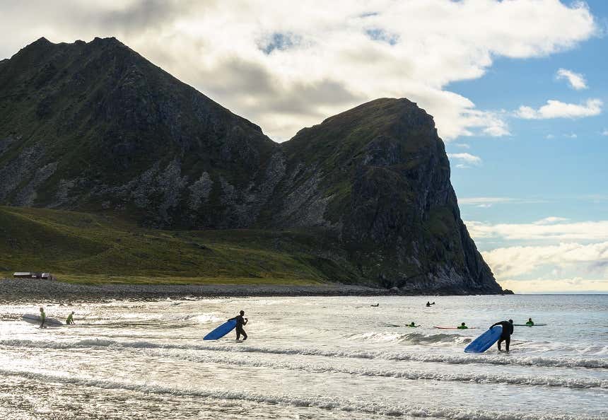 Surfistas entrando no mar na praia de Unstad com uma montanha verde de fundo