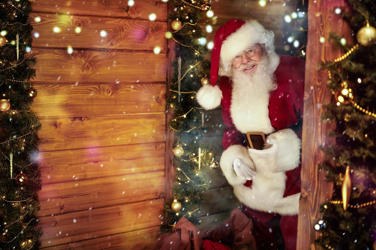 Un hombre vestido de Papá Noel asomándose a la puerta de una casa de madera, decorada con adornos de Navidad.
