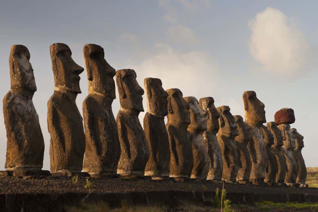 Moais on Easter Island lined up one by one, surrounded by green grass and a blue sky with some grey clouds