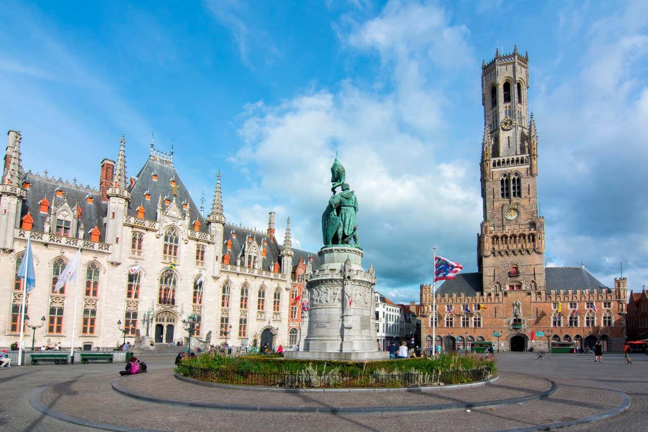 A Grote Markt com um monumento ao centro e um edifício ao lado onde se erguem bandeiras e ao fundo o campanário de Belfort.