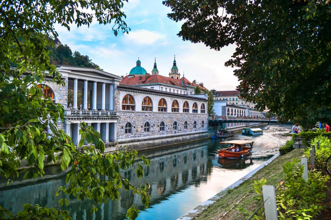 Um barco vermelho atravessando o rio junto aos antigos palácios do centro histórico de Ljubljana, uma das capitais europeias que merece ser visitada