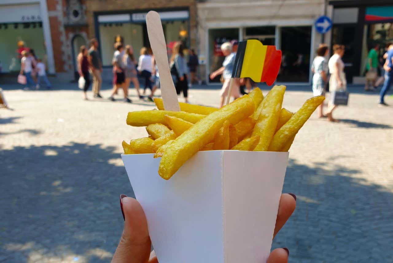 Uma mão segurando uma embalagem de papel com batatas fritas e uma bandeirinha da Bélgica