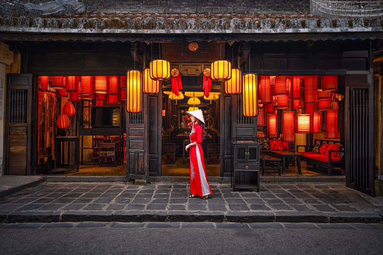 A woman in a traditional red dress walks past an old shop in the centre of Hoi An filled with lanterns.