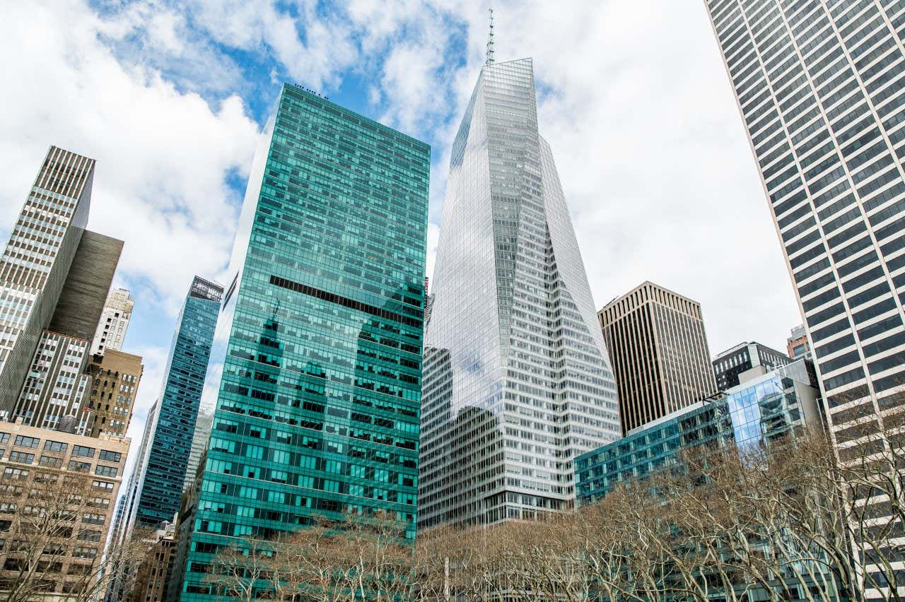 Bank of America Tower seen from the ground alongside other skyscrapers, with a cloudy sky seen behind
