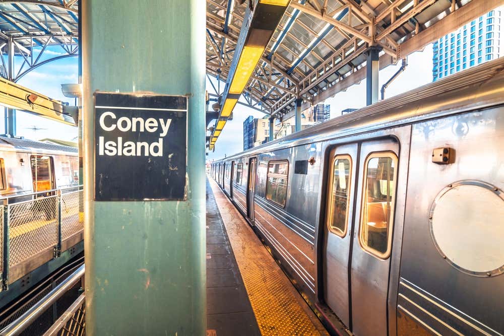Stazione della metropolitana di Coney Island a New York, con cartello nero 'Coney Island' su una colonna verde in primo piano. Treno blu della metro fermo lungo la banchina, sotto una grande tettoia metallica con travi a vista