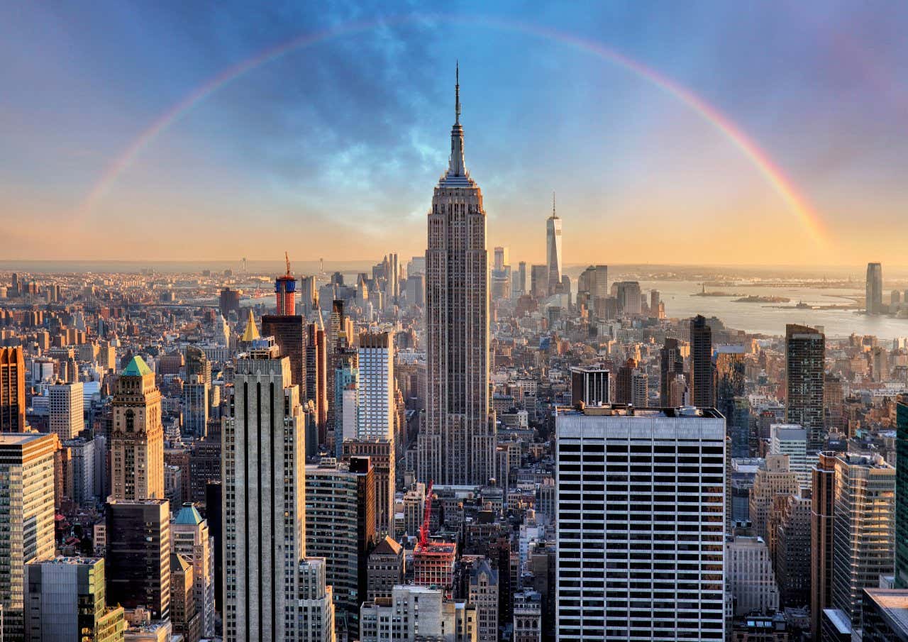 An aerial view of the Empire State Building with more of NYCs skyline in the background as well as a rainbow going over the tower