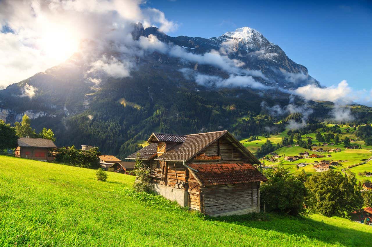 A typical Alpine chalet in the greenery of the pastures, with the mountains in the background, in front of which the clouds hide the sun a little