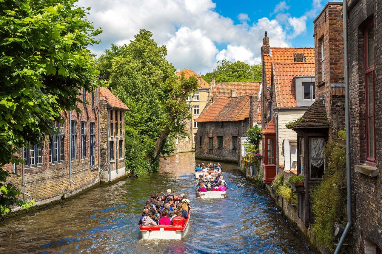 Pessoas passando de barco pelos canais de Bruges rodeados de edifícios típicos 