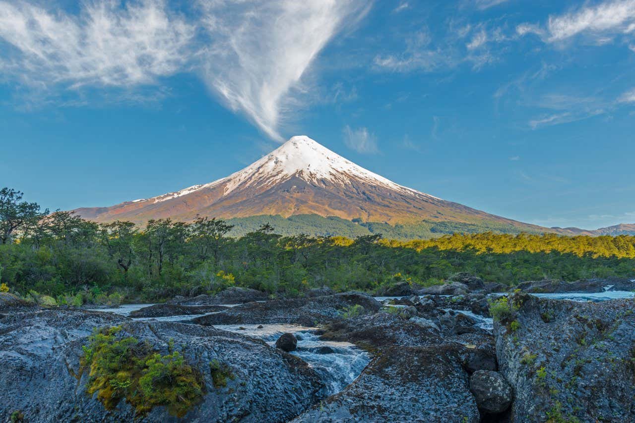 A pointed volcano partially covered in snow under a bright blue sky and some wispy clouds, a verdant lush valley below with flowing rivers and streams