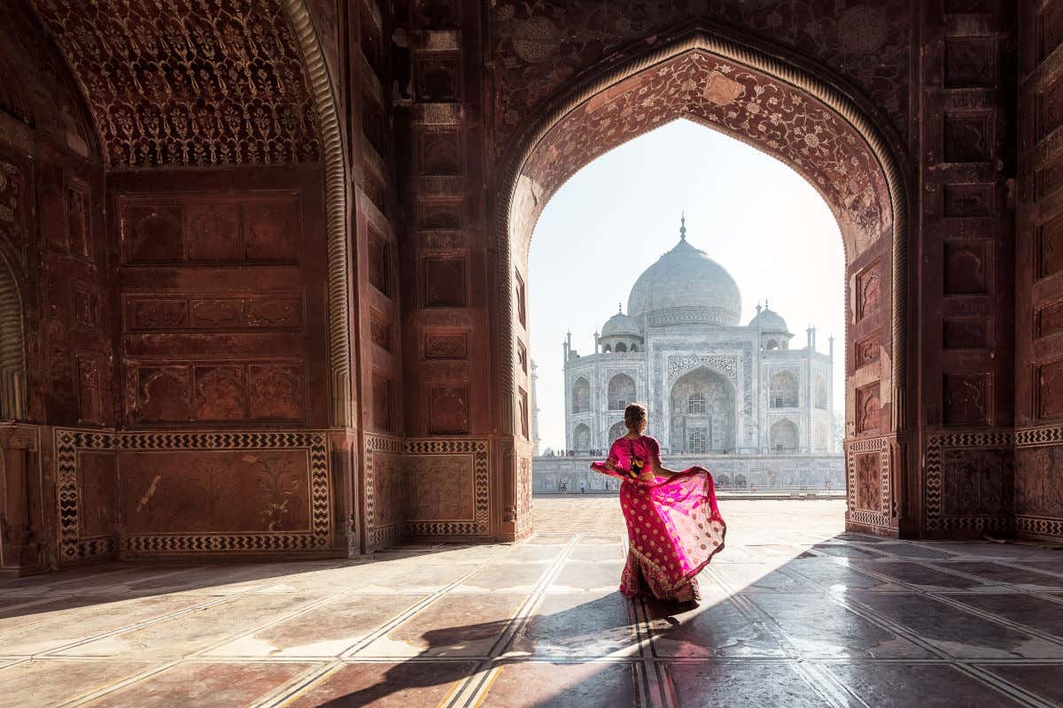Una mujer ataviada con un vestido típico de la India, bailando frente a una puerta de enormes dimensiones con vistas al Taj Mahal