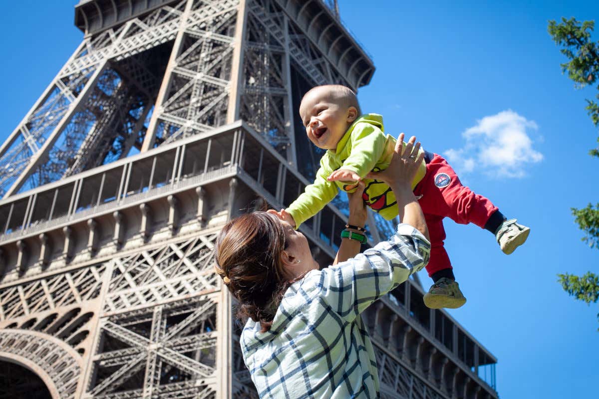 A woman holding a baby in her arms in front of the Eiffel Tower, a clear vibrant blue sky behind the tower, one of the top things to do in paris with kids