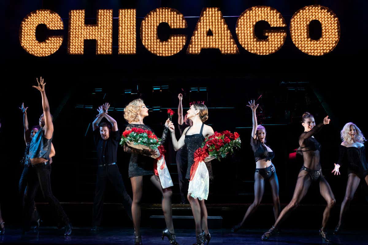 Dos actrices intercambiando rosas frente a un grupo de bailarines del show de Chicago