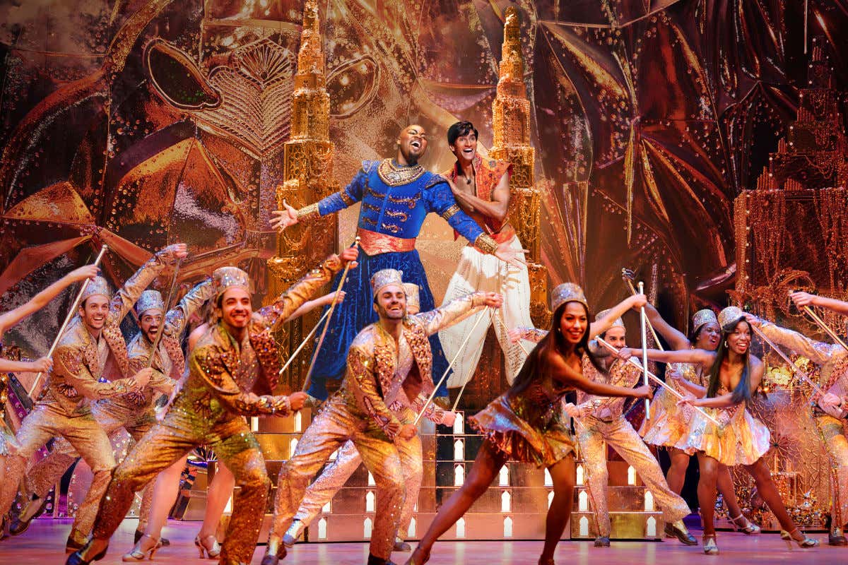 Bailarines con trajes dorados frente a dos actores que interpretan a Aladdín y el Genio en el musical de Nueva York