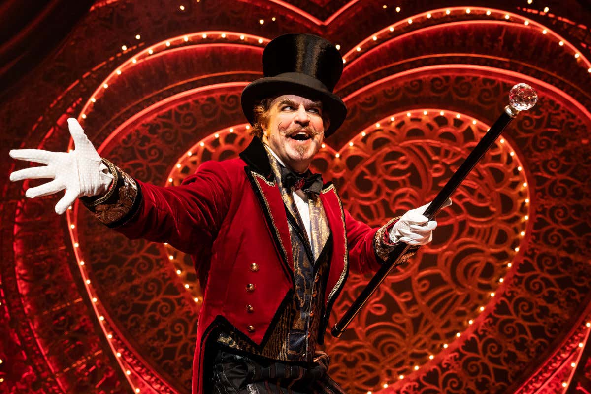 Un hombre ataviado con ropa circense y un bastón frente a un corazón del musical de Moulin Rouge