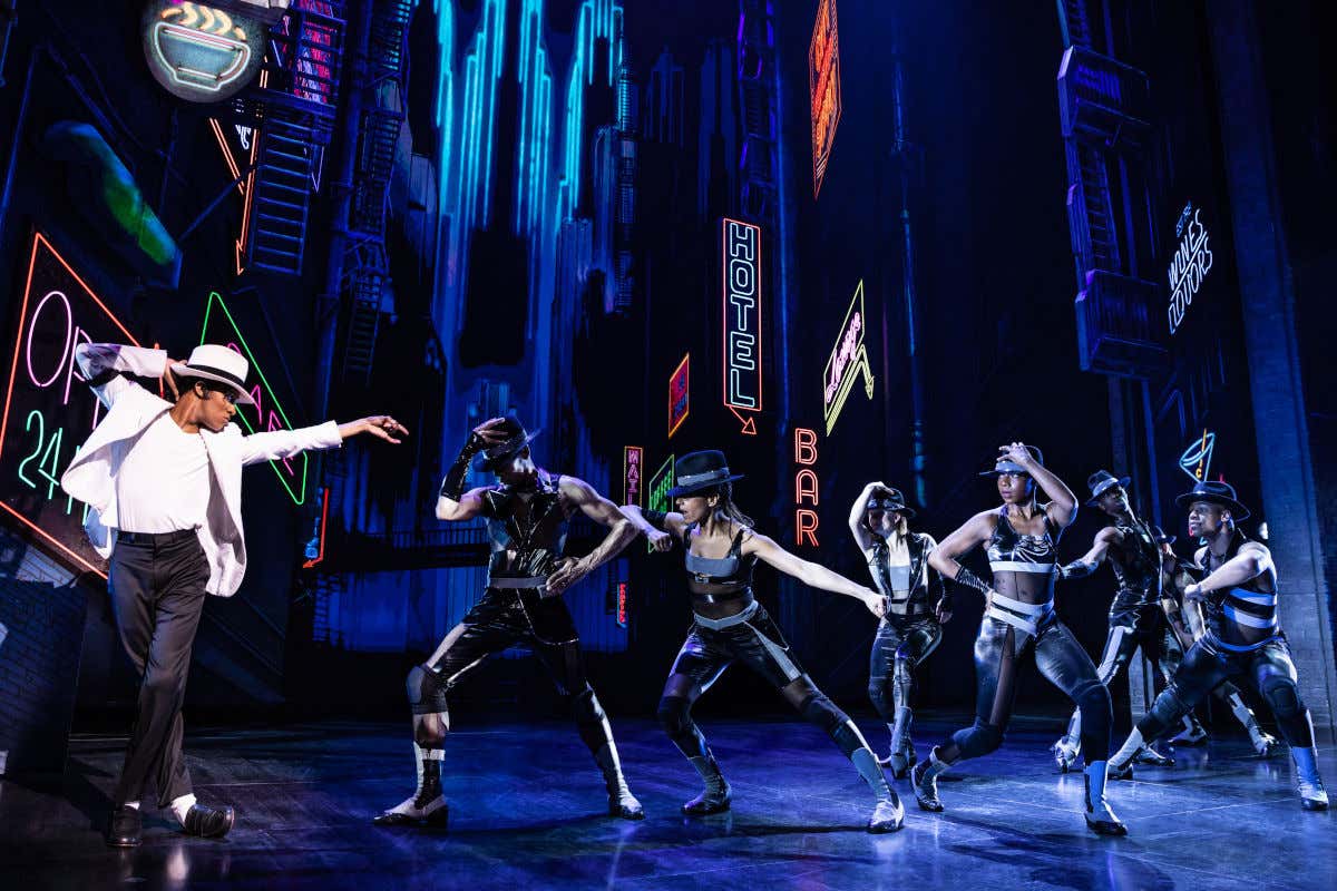 Un actor bailando al estilo de Michael Jackson frente a otros bailarines en un teatro decorado con luces de neón