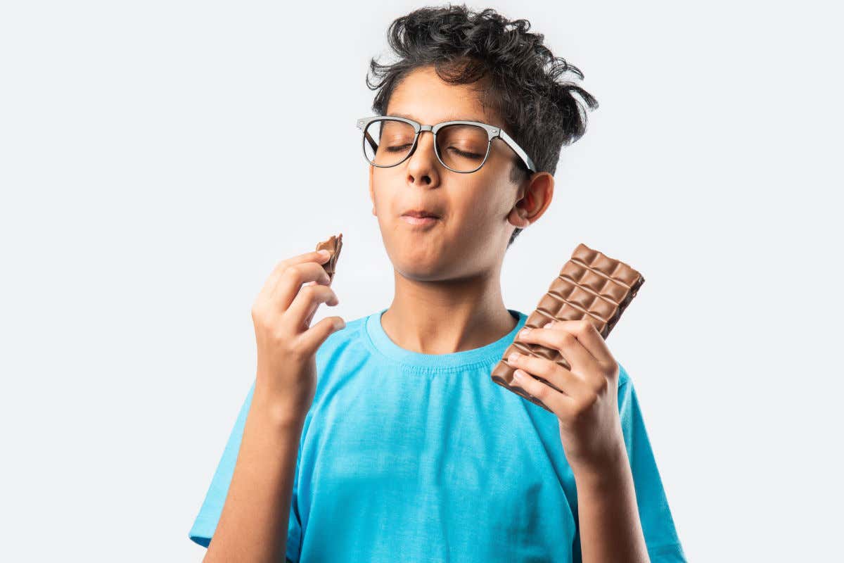 A boy wearing a blue shirt and glasses closes his eyes while enjoying a chocolate bar.