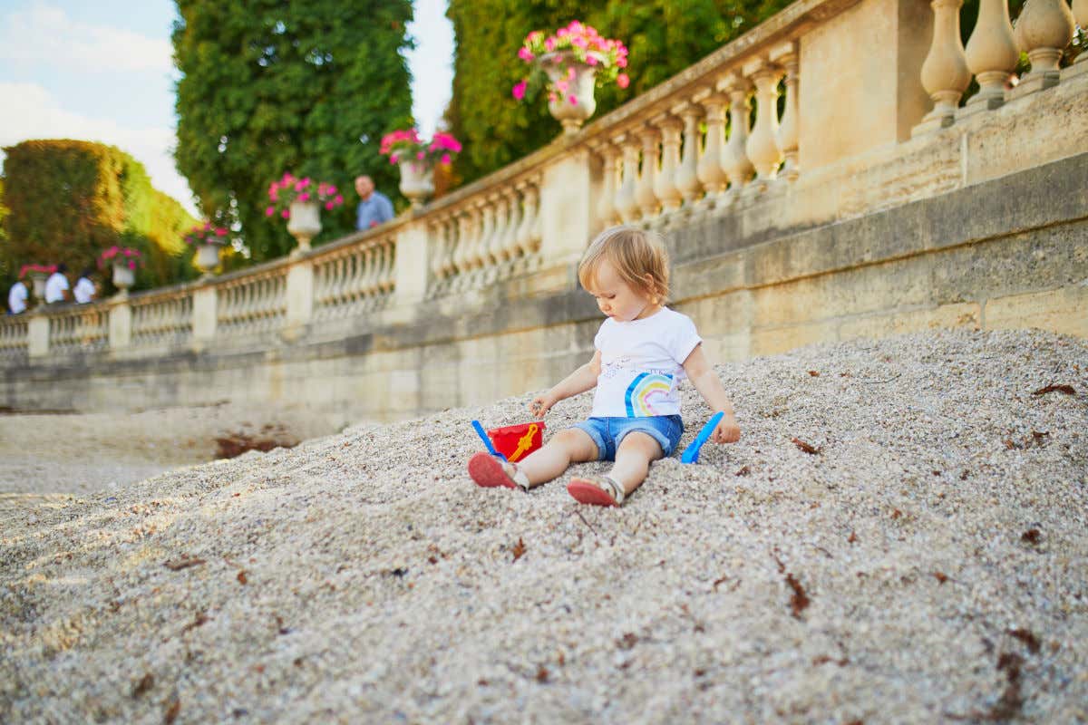 A girl of about two years old sitting on a sand mound in a park in Paris and playing with a bucket and a small shovel