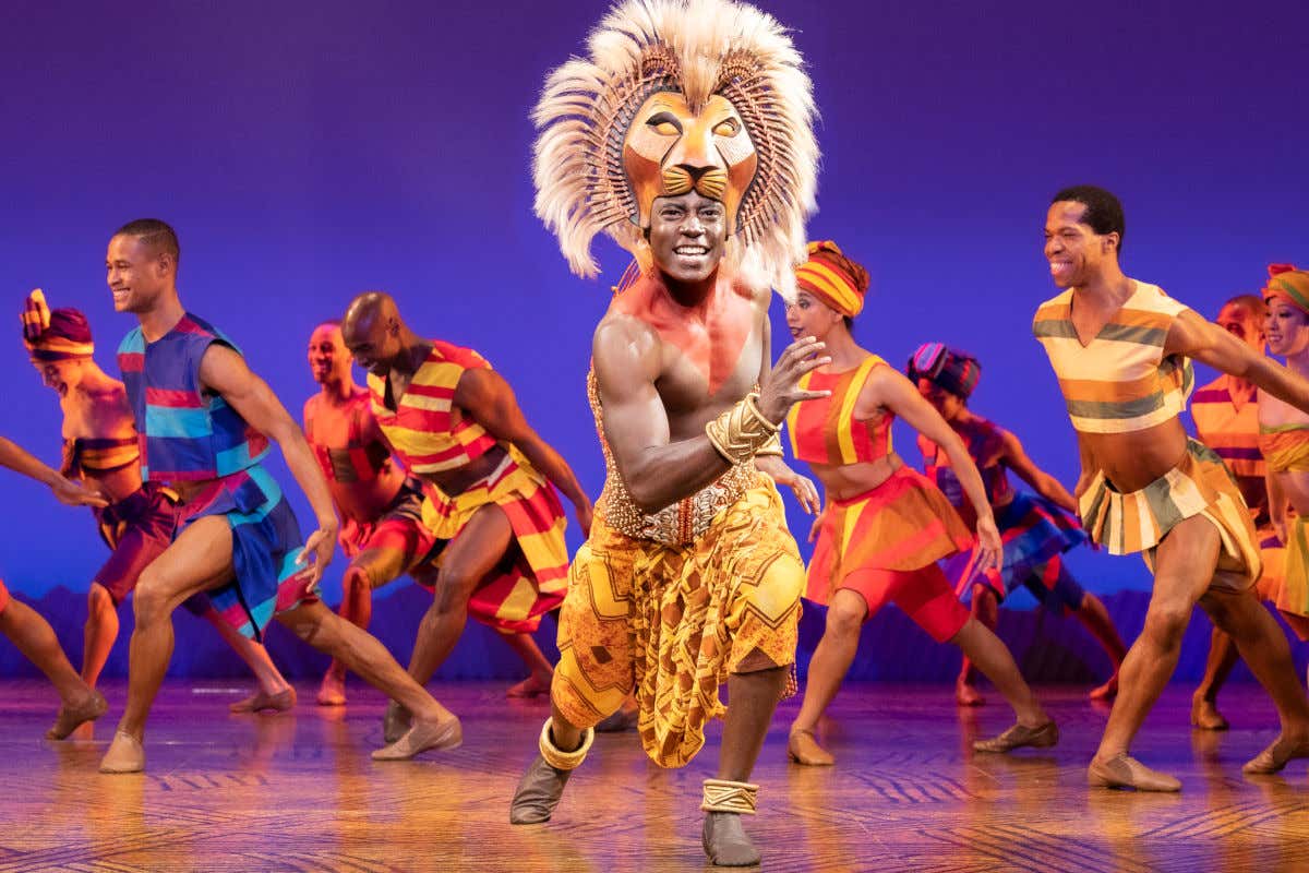 Un actor con una máscara de león y numerosos bailarines con trajes étnicos subidos en un escenario de Broadway representando un musical de Nueva York