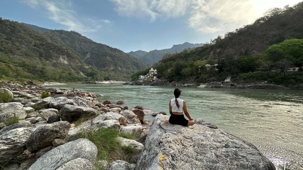 A tourist sits on a rock by a flowing river in Rishikesh, which is surrounded by steep hills covered in vegetation.