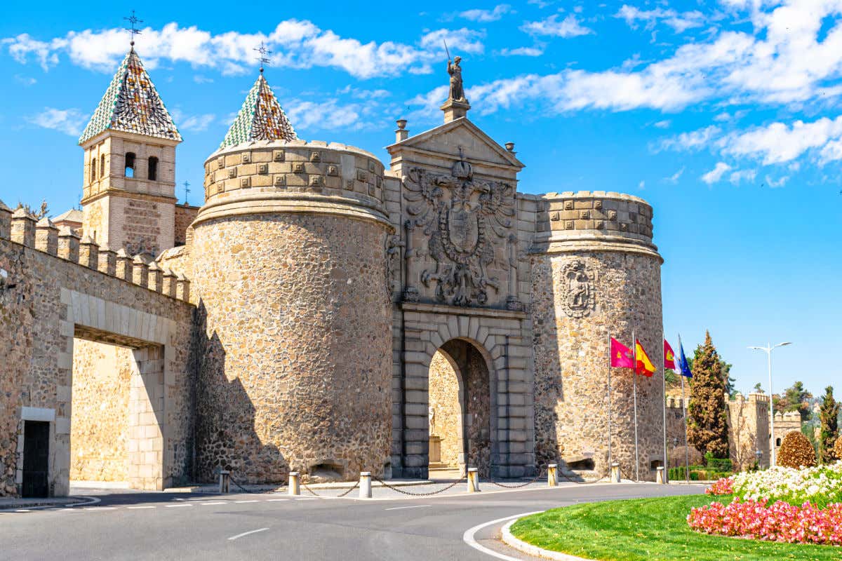 La Puerta de Bisagra, un pórtico monumental con un enorme escudo de piedra frente a una rotonda y varias banderas