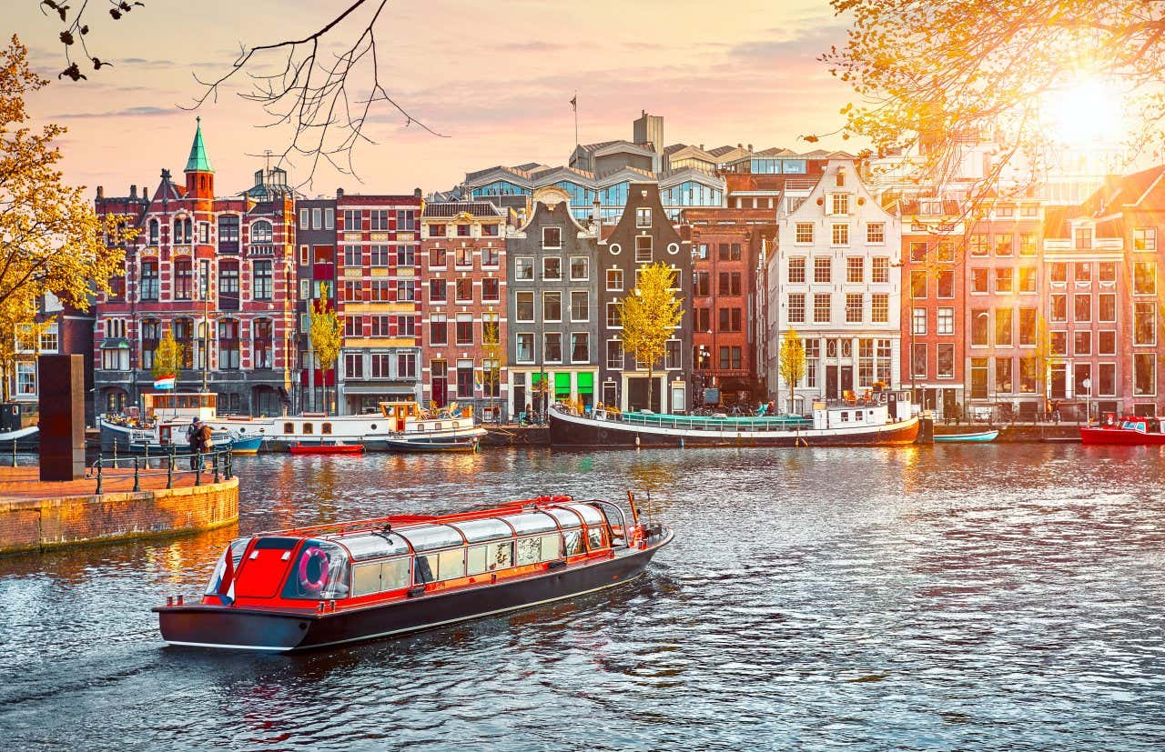 Bateau naviguant au premier plan sur un canal d'Amsterdam et maisons aux façades colorées en arrière-plan sous un ciel dégagé avec le soleil en face