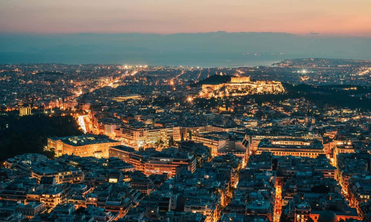 An aerial shot of Athens lit up at night, with the Athens Acropolis visible in the distance.