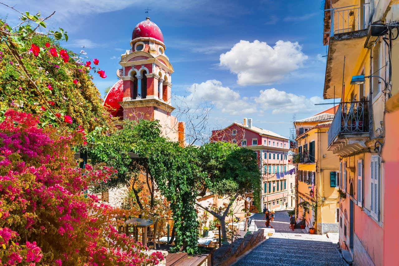 Una strada piena di fiori e case colorate, con una chiesa dall'architettura tipica, sotto il cielo azzurro dell'isola greca di Corfù