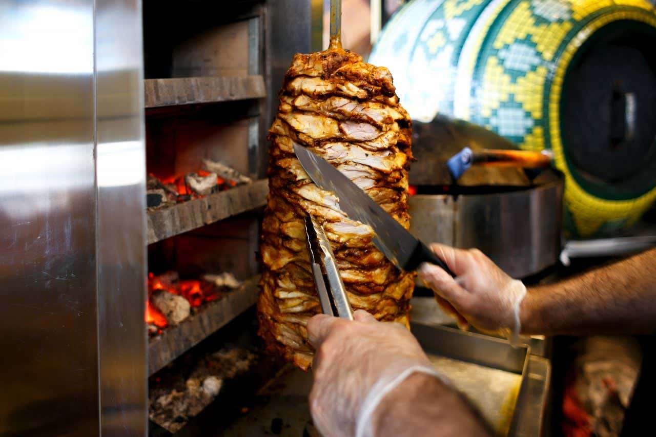 A chef wearing plastic gloves cuts the meat from meat cooked on a vertical rotating spit.