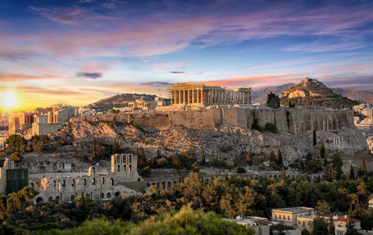 The Acropolis of Athens, with the Parthenon in the centre, as seen from afar, with the sun setting in the background on the left side of the image.