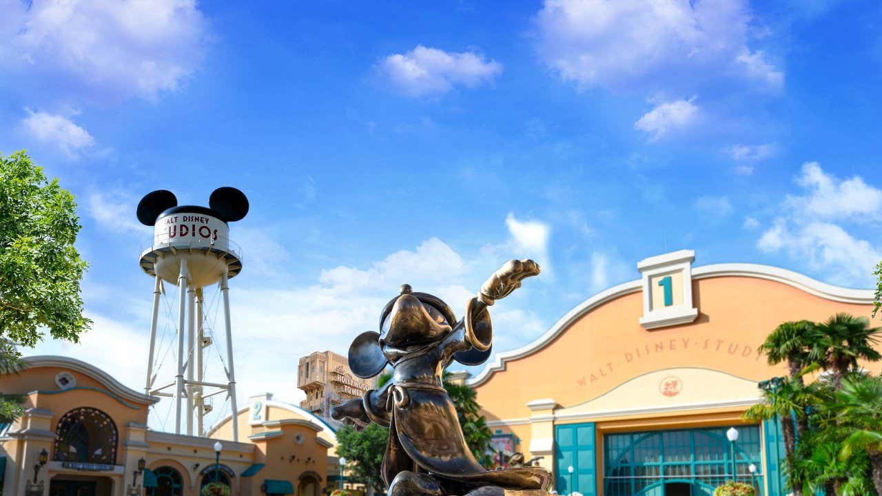 A Mickey Mouse statue facing the camera, with Disney studio buildings and a cloudy sky in the background.