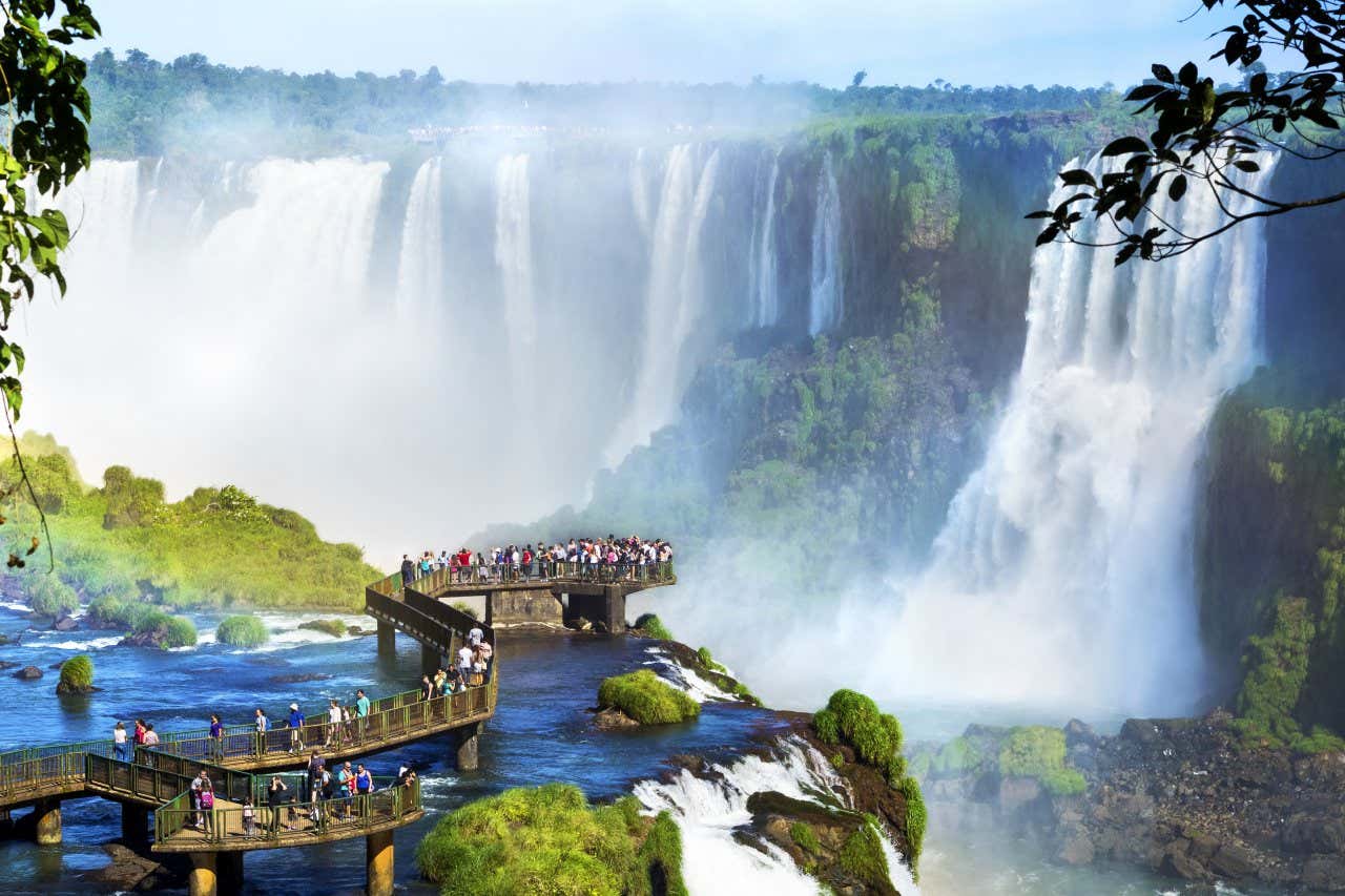 Turistas passeando pela passarela sobre as Cataratas do Iguaçu e a paisagem natural das quedas d'água ao fundo
