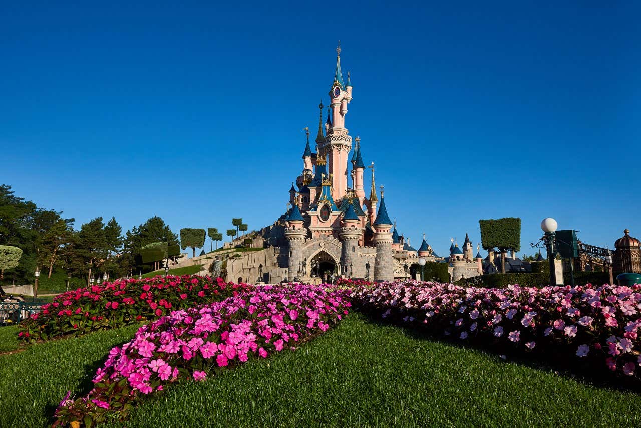 A wide shot of Sleeping Beauty's Castle at Disneyland Paris, with a clear blue sky in the backgrouund, and colourful flowers in the foreground.