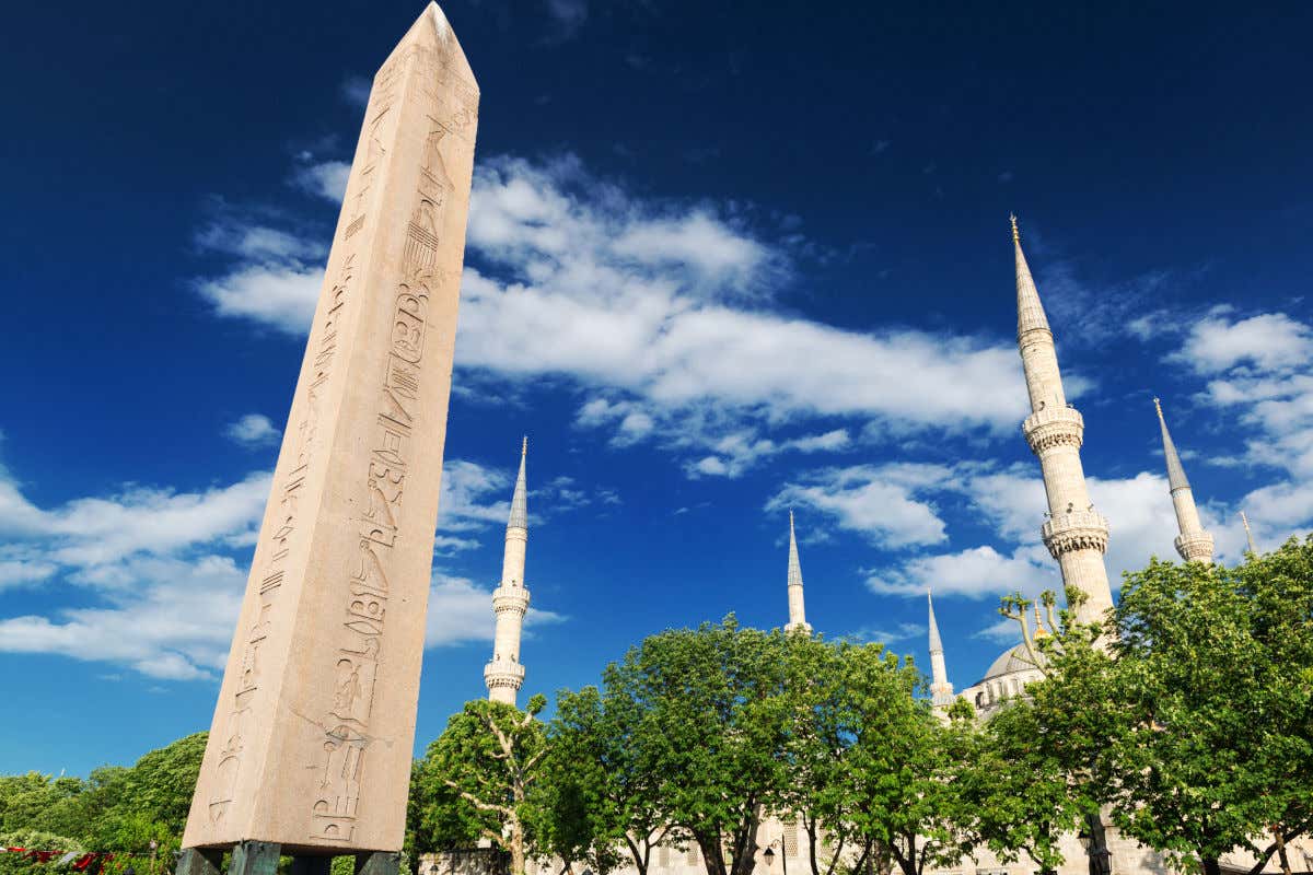 Un obelisco con jeroglíficos egipcios junto a varios árboles y las torres de una mezquita
