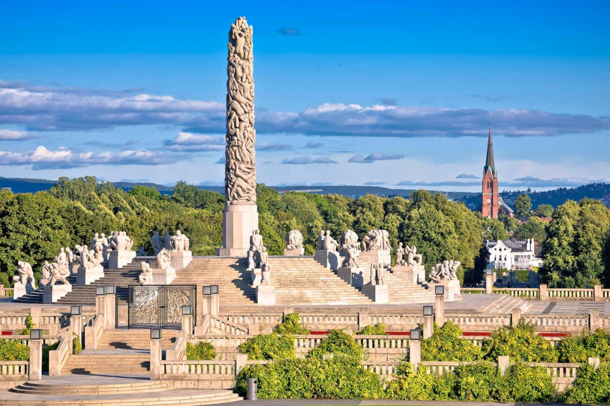 Una enorme columna repleta de esculturas con formas humanas en mitad de un parque con vistas a una iglesia de ladrillo rojo