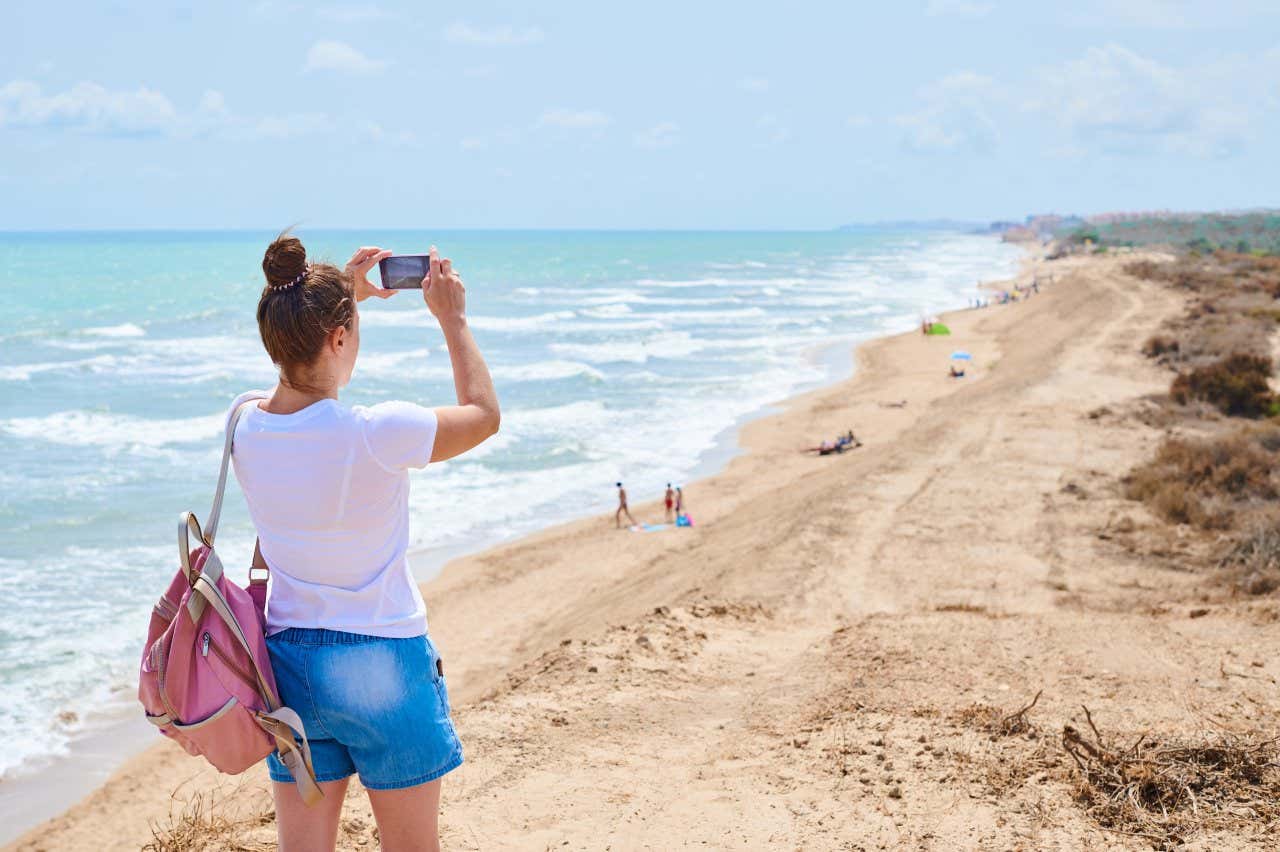 Una ragazza di spalle scatta una foto a una lunga spiaggia assolata e ventosa