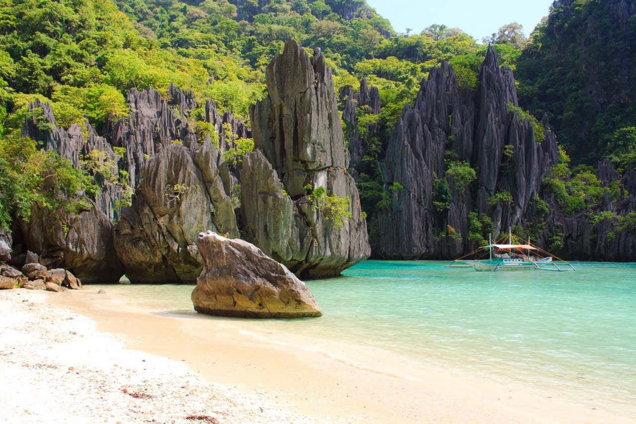 Una spiaggia tropicale di sabbia bianca con formazioni rocciose verticali e una piccola barca di legno in acqua
