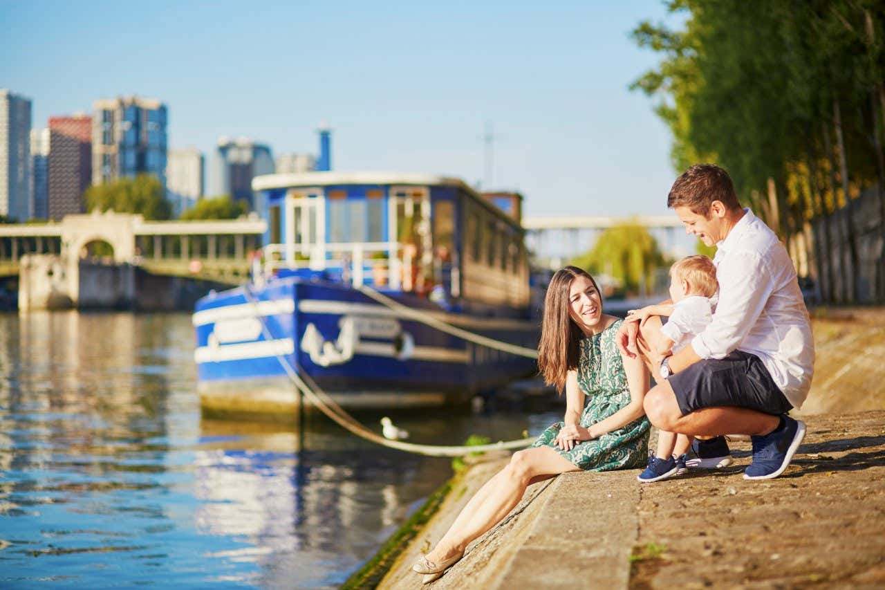 A family of three sit by the edge of the River Seine, a blue boat behind them out of focus, the Paris skyline even farther in the distance