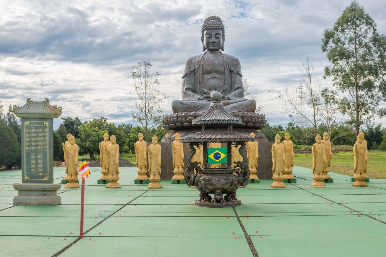 Diversas estátuas amarelas em frente de uma grande estátua de metal. No plano da frente, um grande incensório com a bandeira do Brasil