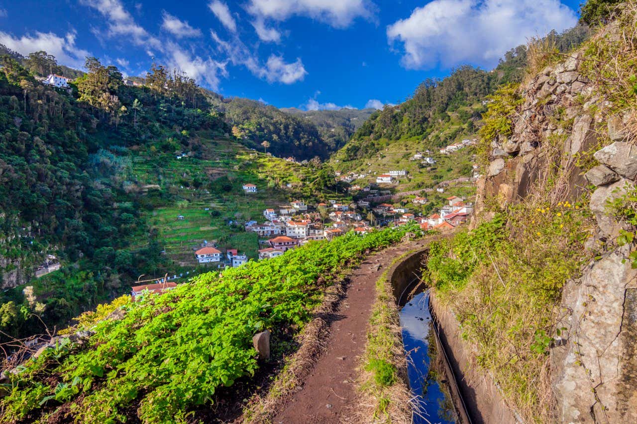 A view of a levada running around a mountain with a village of white houses in the valley on a sunny day.
