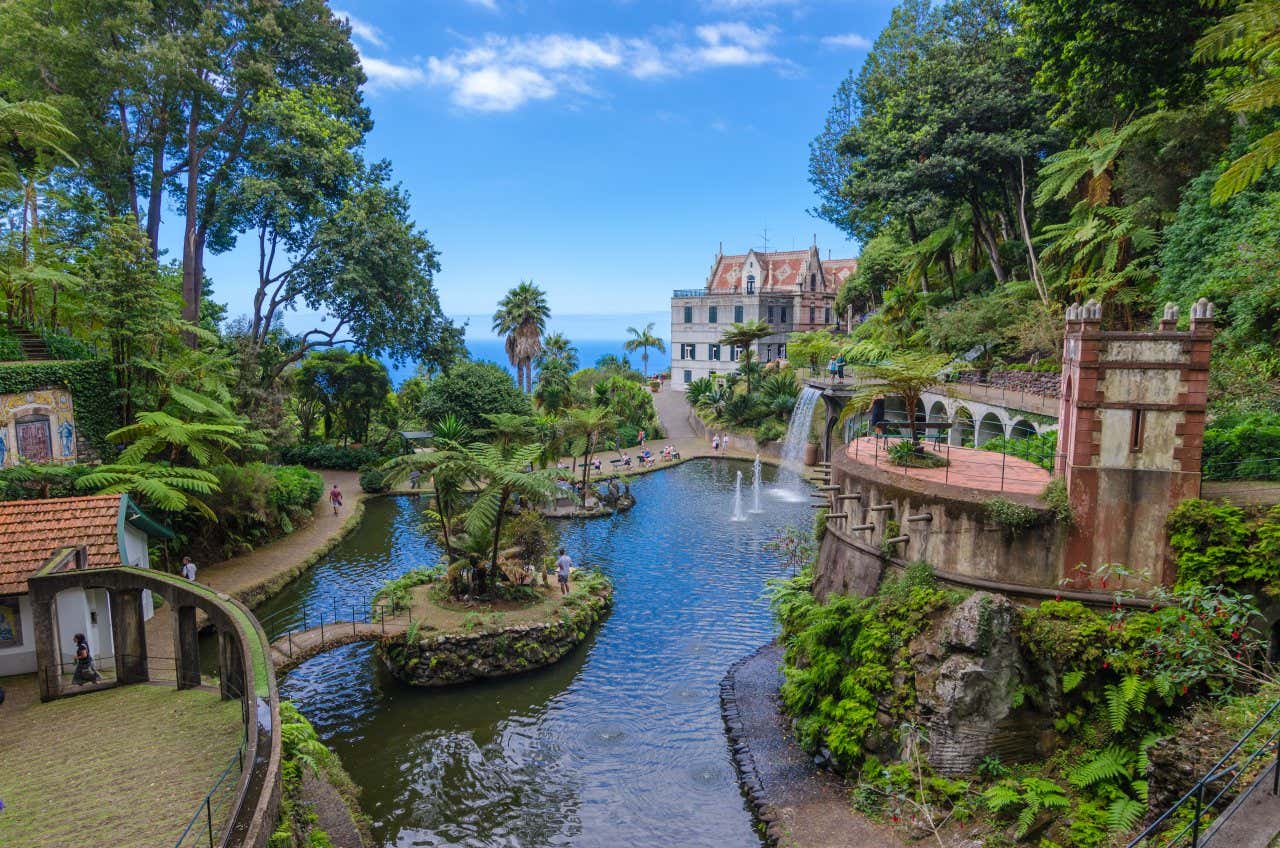 A view of the Monte Palace Gardens, with a pond surrounded by green trees and people walking along the paths on a sunny day.