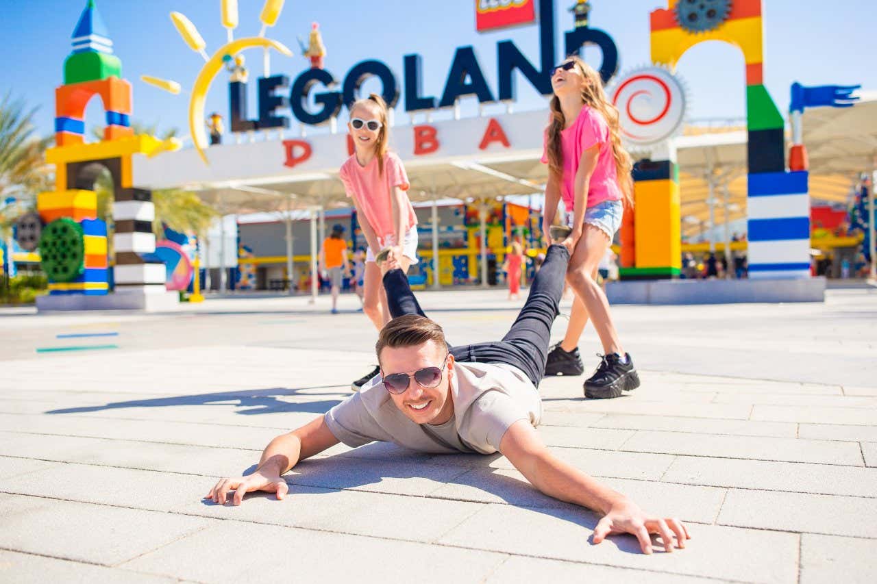 Two girls in pink t-shirts and denim shorts pulling the legs of a man lying on his front outside LegoLand Dubai