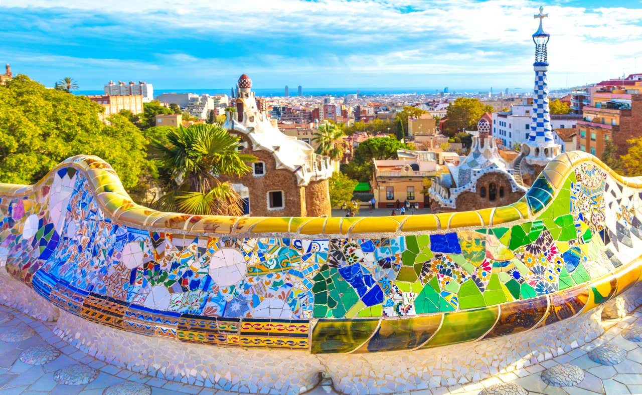 A low wall decorated in colourful mosaics overlooking stone buildings with mosaic-tiled roofs and the city of Barcelona