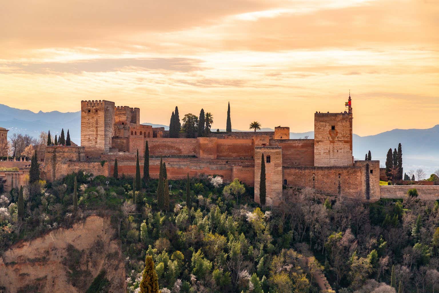 Vista sul complesso dell'Alhambra circondata da alberi e vegetazione al tramonto e con delle montagne sullo sfondo